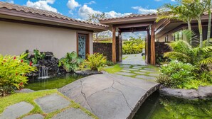 L’entrée d’un complexe hôtelier tropical, avec un chemin de pierre menant à une zone délimitée par une grille, entourée d’une végétation luxuriante et d’une petite cascade.