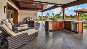 Une cuisine extérieure moderne dotée d’un îlot intégré, d’un coin salon avec des meubles en osier et offrant une vue sur un paysage tropical.