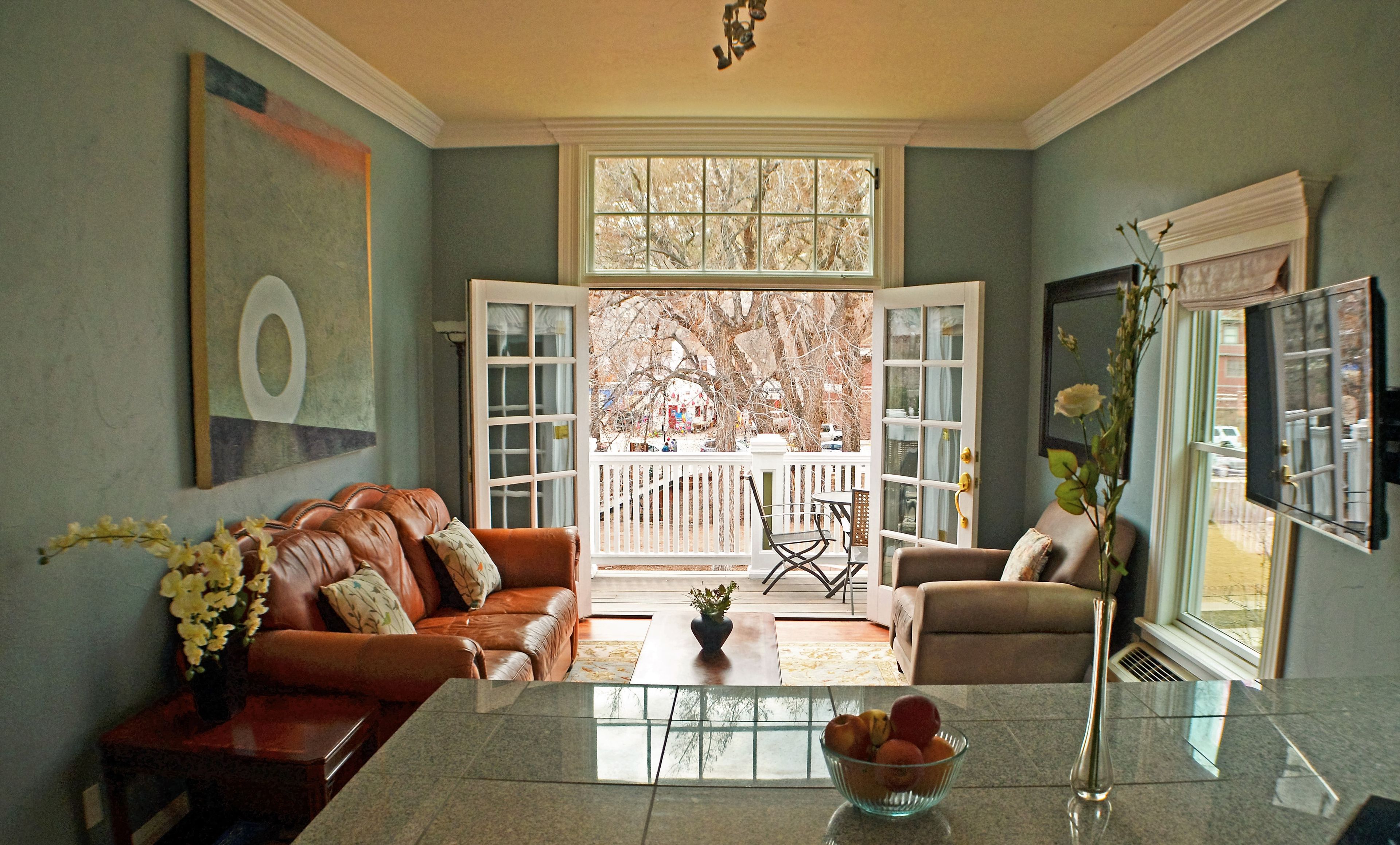 A spacious living room with a large window, a brown leather sofa, a glass coffee table, and a TV on the right.