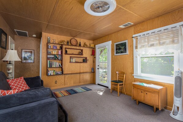 A cozy room with a wooden ceiling, a bookshelf, a window with curtains, a fan, and a couch with pillows. - River Oasis with Plenty of Family Fun (Three Rivers)