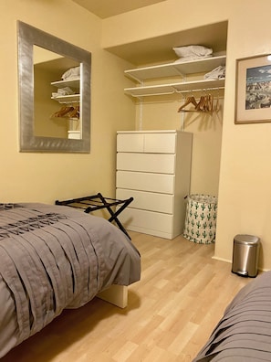 A bedroom with a bed, a dresser, a mirror, a closet with shelves, and a trash can.