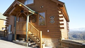 Une cabane en bois, construite sur des fondations en pierre, dotée d’un porche avec une balustrade en bois et d’un escalier menant à l’entrée.