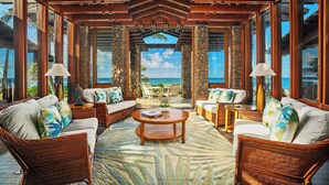 Una habitación amplia con muebles de mimbre, una mesa de centro y ventanas grandes con vista al océano.