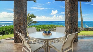 A round outdoor table with four chairs, a wooden tray with two blue mugs, and a view of the ocean and beach.