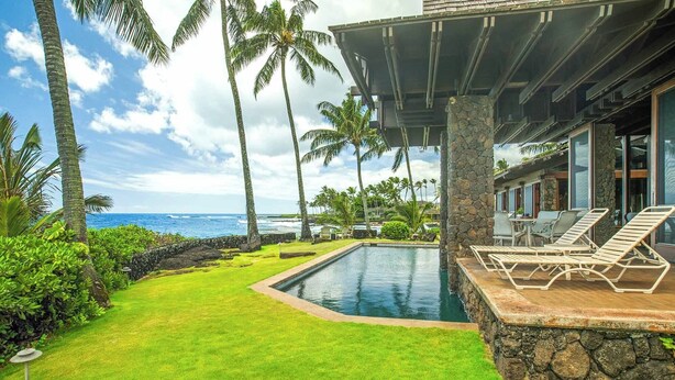 A pool area with lounge chairs, a stone wall, and a view of the ocean.