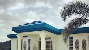 A two-story building with a blue roof and white walls. There is a covered porch with white columns and a white chair. The building has decorative windows and a small garden area with a palm tree. - Casa Menta in Rincon, Puerto Rico (Rincon)