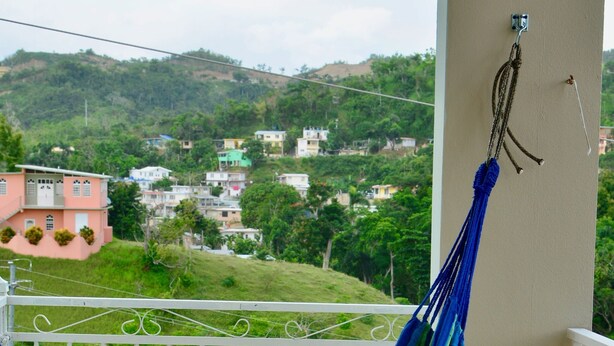 A balcony with a blue hammock, a chair, and a view of houses and greenery in the background. - Casa Menta in Rincon, Puerto Rico (Rincon)
