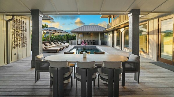 A covered outdoor area with a pool, lounge chairs, and a dining table.