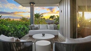 Un área de estar al aire libre techada con un sofá y sillones, una mesa redonda y vistas al atardecer sobre palmeras.