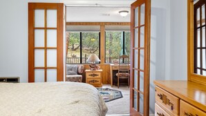 Un dormitorio con una cama grande, cómoda de madera y un escritorio con silla. Amplias ventanas ofrecen vistas a árboles y un balcón.