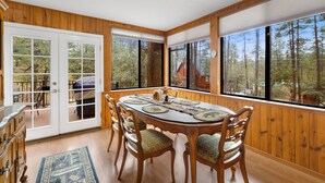 Comedor con mesa de madera, sillas y vistas a árboles a través de amplias ventanas.