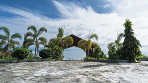 A stone-paved area with a wooden archway, surrounded by palm trees and greenery, leading to a body of water. - Estrella Hotel & Conference (Luwuk)