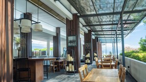 A modern restaurant with wooden tables and chairs, a bar area, and a view of the sea. - Estrella Hotel & Conference (Luwuk)
