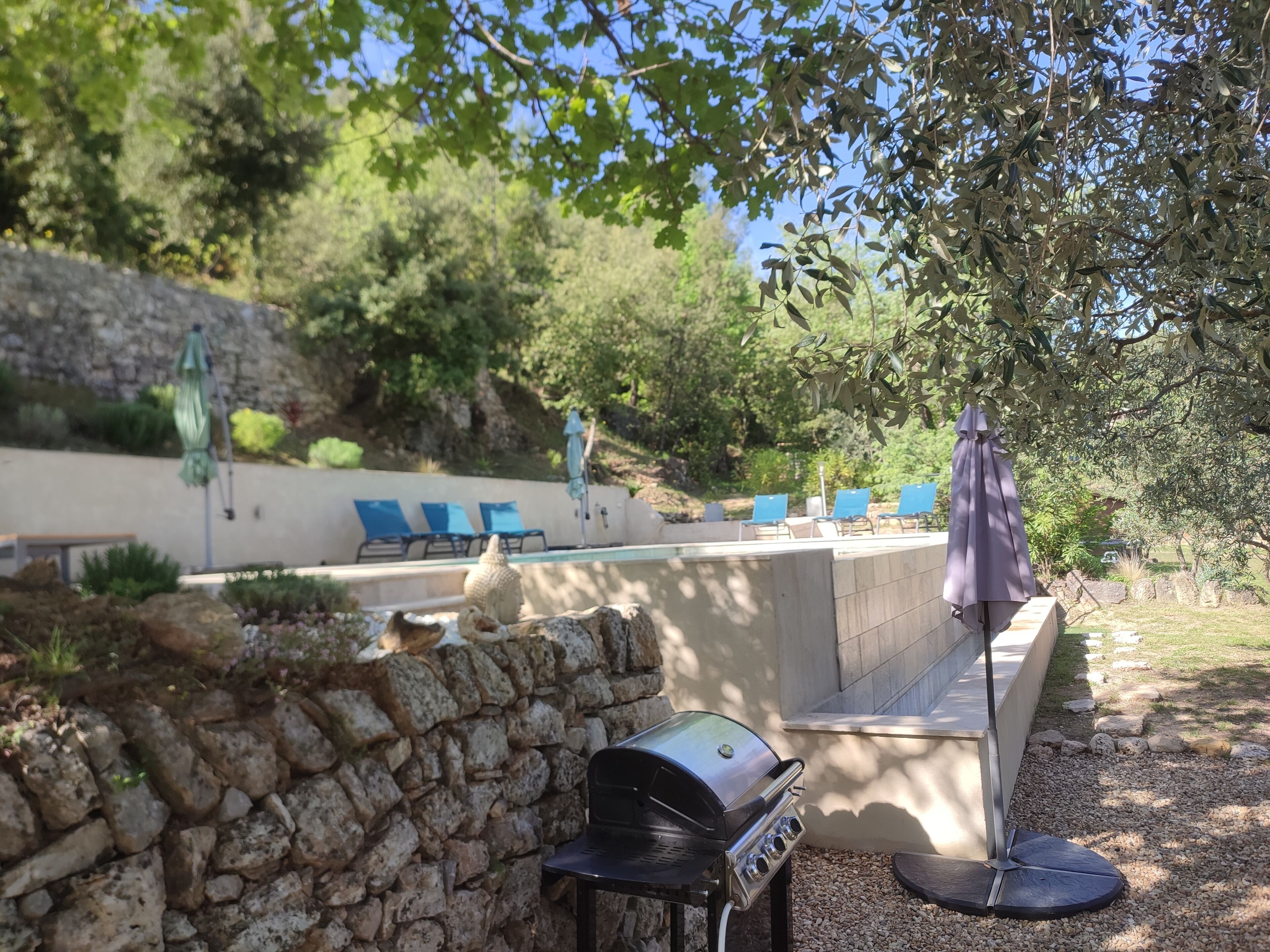 A stone wall, a barbecue grill, and blue lounge chairs by a pool.