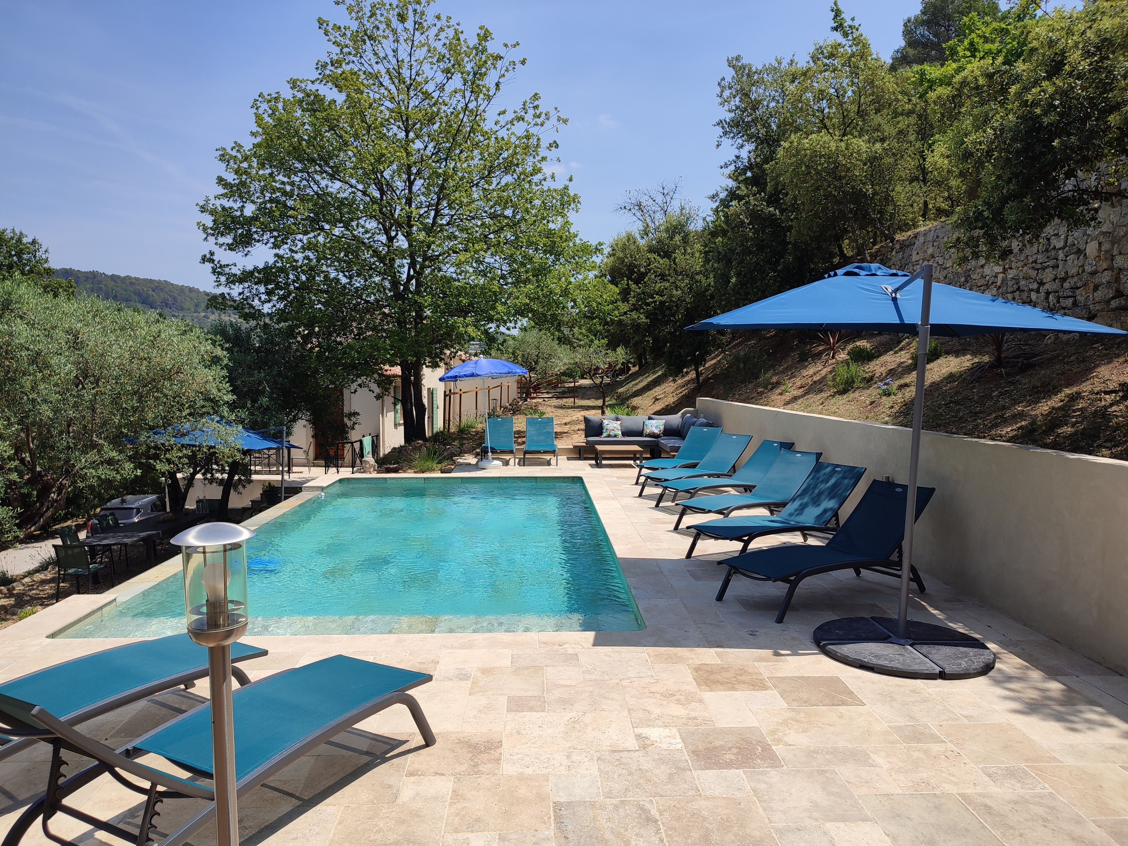 Un espace piscine agrémenté de chaises longues et de parasols, entouré d’arbres et d’un mur de pierre.