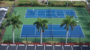 Ein Tennisplatz mit zwei Spielern, umgeben von Palmen und einem Parkplatz.