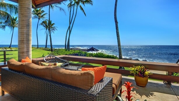 A wicker sofa with orange cushions, a small table, and a potted plant on a balcony overlooking the ocean with palm trees and a view of the sea. - Hale Makai By Parrish Kauai- oceanfront with pool and AC, honeymoon haven at Hal (Koloa)