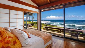 A bedroom with a large window overlooking the ocean, a bed with patterned bedding, and a view of the beach and rocky shoreline. - Hale Makai By Parrish Kauai- oceanfront with pool and AC, honeymoon haven at Hal (Koloa)