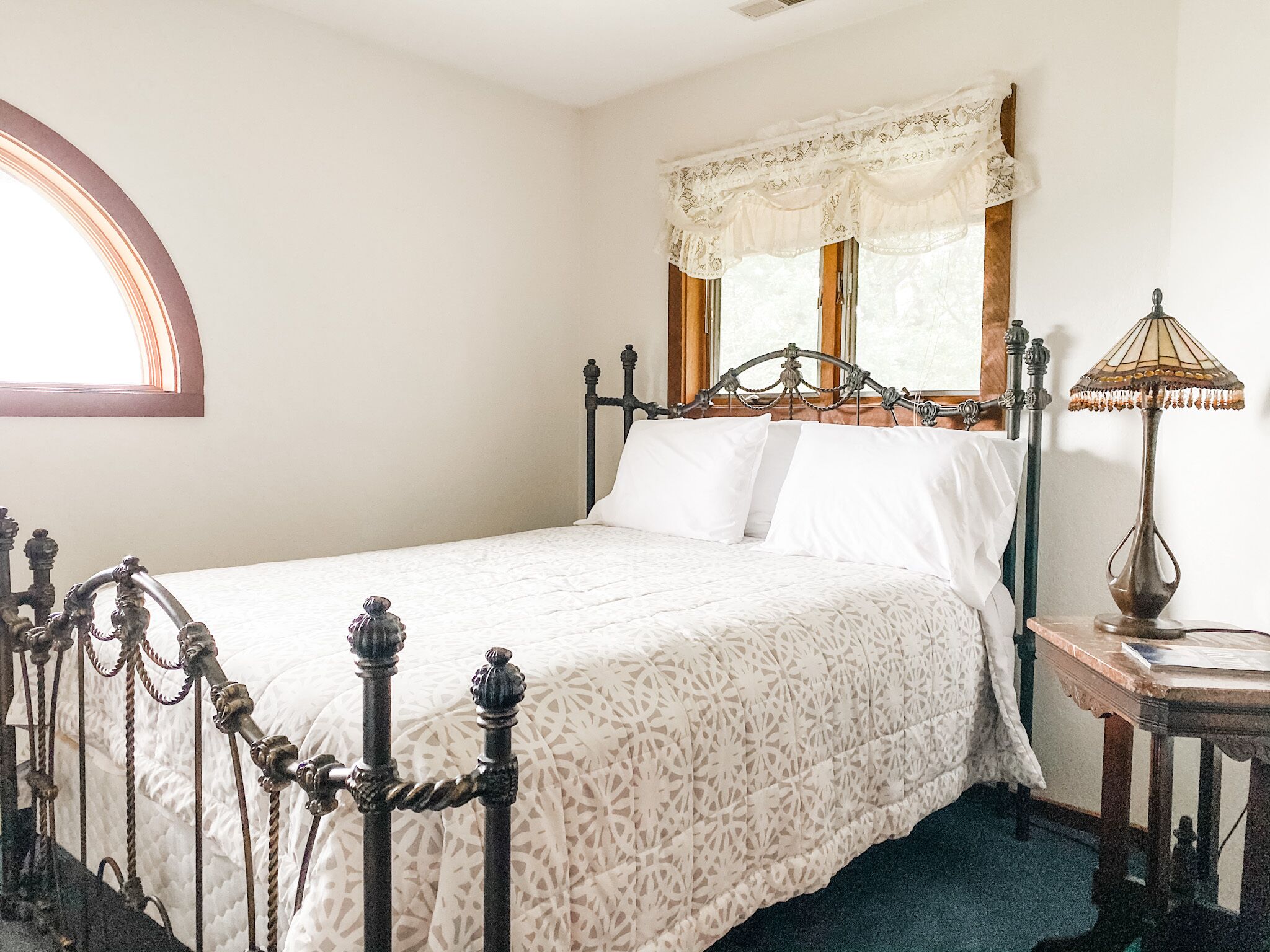 A bedroom with a four-poster bed, a nightstand, a lamp, and a window with lace curtains.