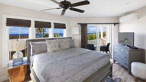 Une chambre avec un lit, un ventilateur de plafond, une télévision, une commode et une vue sur l’océan grâce à de grandes fenêtres.