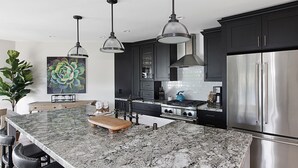 Une cuisine moderne avec des meubles de couleur sombre, un grand îlot central doté d’un plan de travail en marbre, des appareils en acier inoxydable, des luminaires suspendus et une grande plante verte.