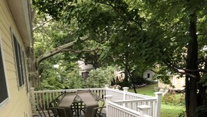A wooden deck with a white railing, a table and chairs, and a view of a green yard with trees and a house in the background. - Cozy cottage in Sandy Pond (Pulaski)