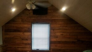 A room with wooden panel walls, a sloped ceiling, a ceiling fan, a window with blinds, and a bed with green bedding. - Cozy cottage in Sandy Pond (Pulaski)