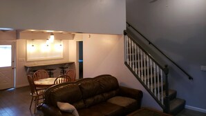 A living room with a brown leather sofa, a dining table with chairs, and a kitchen area in the background. There is a staircase with a black railing leading to an upper level. - Cozy cottage in Sandy Pond (Pulaski)