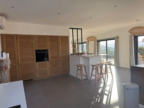 A modern kitchen with wooden cabinets, a central island with stools, and a view through large windows. - Villa breathtaking sea view with pool, 550m from the beach, for 14 people (Serra-Di-Ferro)