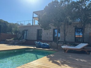A pool area with a wooden deck, lounge chairs, and a stone building with a stone facade and windows. - Villa breathtaking sea view with pool, 550m from the beach, for 14 people (Serra-Di-Ferro)