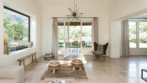 A spacious living room with a large window, a wooden ceiling, and a modern chandelier.