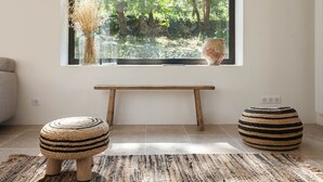 A room with a large window, a wooden bench, two woven stools, and a textured rug.
