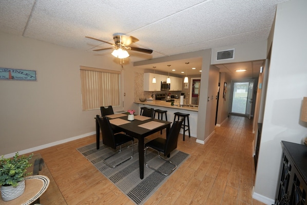 A dining area with a table and chairs, a ceiling fan, and a kitchen in the background.