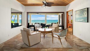 A spacious room with a stone floor, a round table, and chairs, overlooking a beach with the ocean.