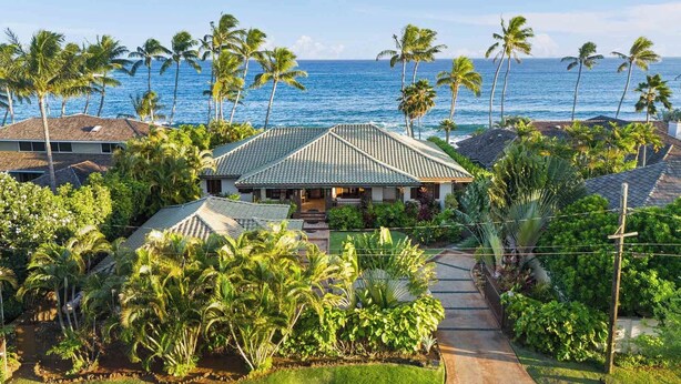 A house with a tiled roof, surrounded by lush greenery and palm trees, overlooking a clear blue ocean.