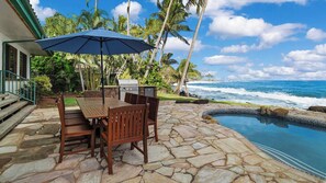 An outdoor patio with a stone floor, a wooden table with chairs, a grill, and a view of the ocean.