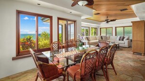 A spacious dining area with a glass table and wicker chairs, large windows with ocean views, a wooden ceiling fan, and a cozy seating area with a sofa and cushions.