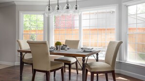 Un comedor con una mesa de madera y cuatro sillas, ventanas amplias con vista a una valla de madera, y lámparas colgantes.