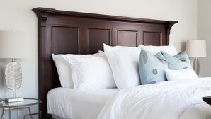 Una cama bien hecha con almohadas blancas y estampadas, un cabecero de madera oscura y una mesita redonda con una lámpara.