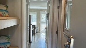 Corredor com uma cama à esquerda, uma porta à direita e vista para um quarto com janela e poltrona.