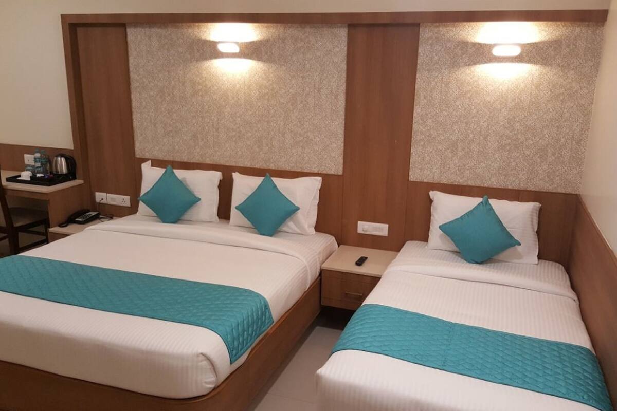 A hotel room with two beds, each with a teal bedspread and white bedding. There is a wooden headboard with a textured design and two wall-mounted lights. A small bedside table with a phone and a remote control is visible.
