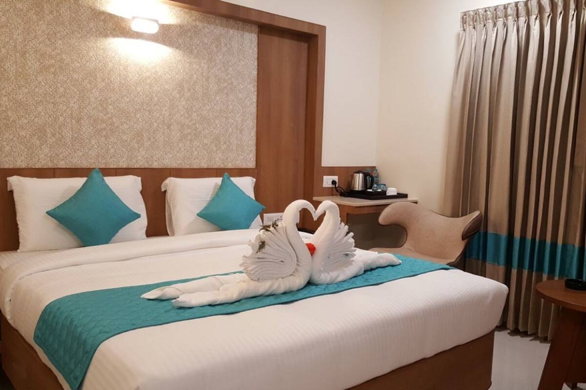 A hotel room with a neatly made bed, a wooden headboard, a small table with a kettle, and a curtain.
