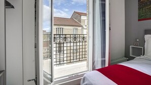 A bedroom with a bed, a nightstand, a window with a view of buildings, and a door. - Paris hôtel (Clichy)