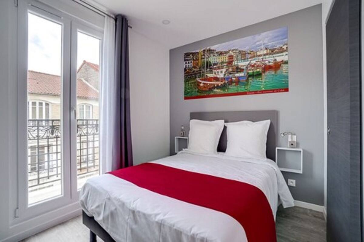 Habitación de hotel con una cama grande, un cuadro de un paisaje urbano en la pared, un balcón con vista a edificios y una ventana con cortinas.