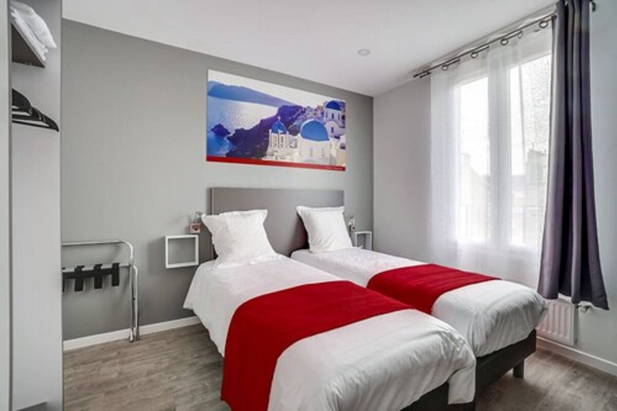 Habitación de hotel con dos camas, una colcha roja y blanca, un cuadro de una escena costera y una ventana con cortinas.