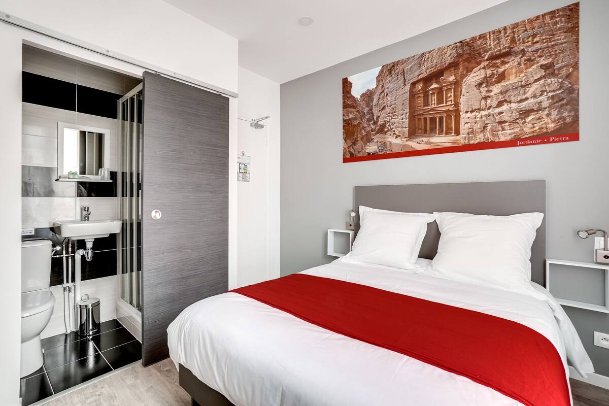 Una habitación de hotel moderna con una cama grande, un detalle rojo, un baño con lavamanos e inodoro, y una obra de arte en la pared que representa Petra.