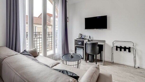 A modern living room with a sofa, a small round table, a TV mounted on the wall, and a window with curtains. - Paris hôtel (Clichy)