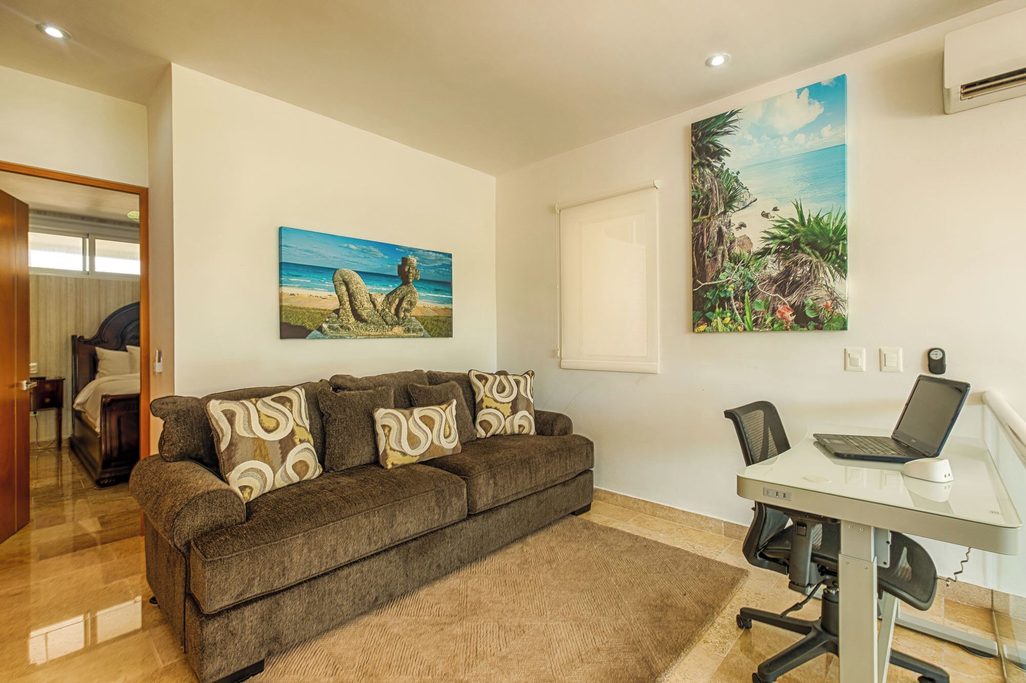 A living room with a sofa, a desk with a laptop, and two paintings on the wall.