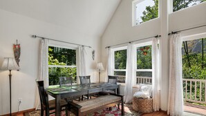A dining area with a table and chairs, a bench, a lamp, and a view of greenery through large windows.