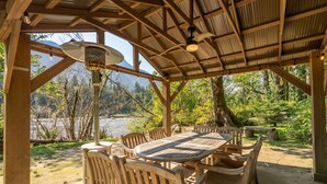A wooden gazebo with a round table and chairs, overlooking a lake and forested area.